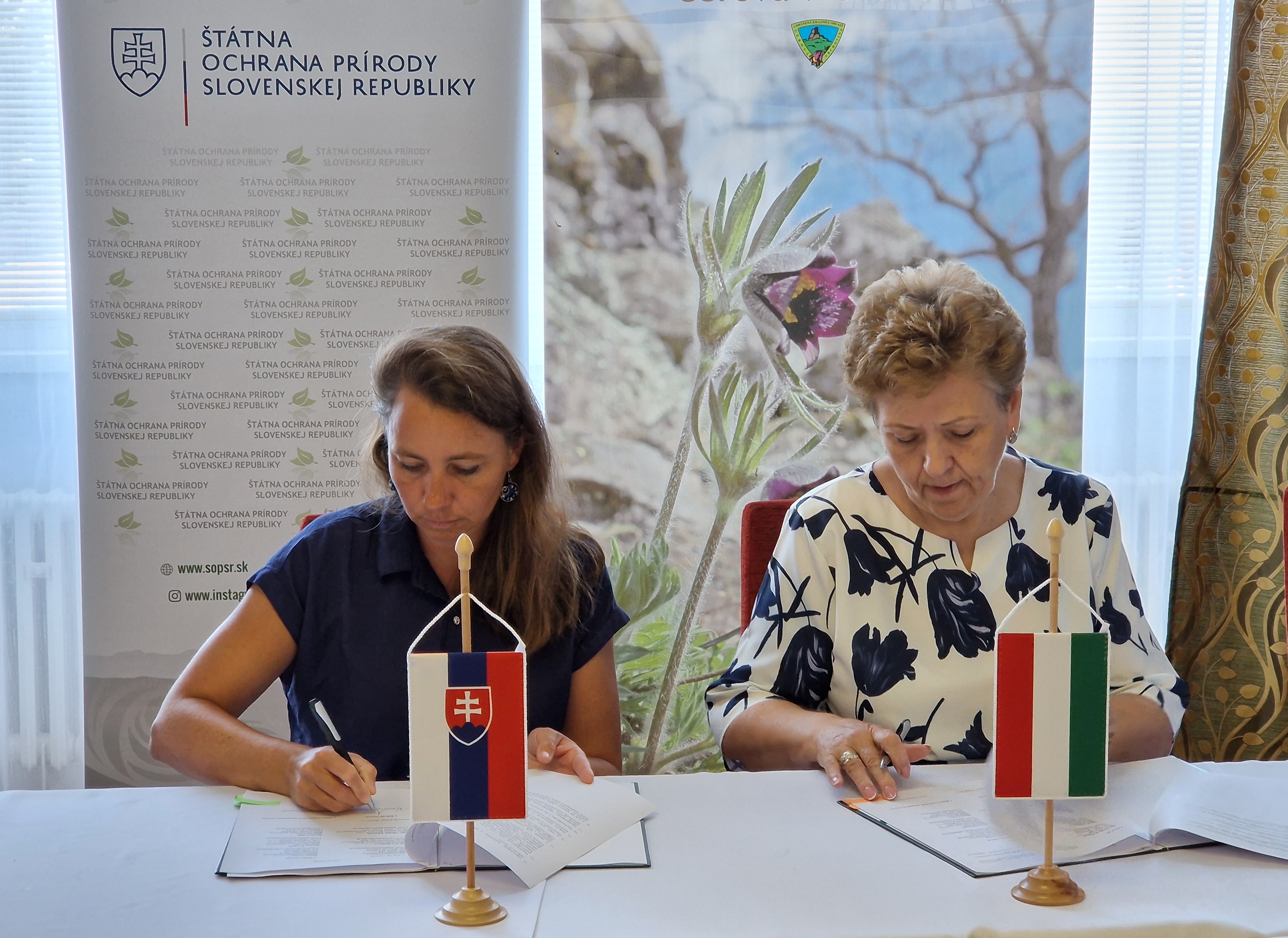 Na obrázku sú dve ženy, každá sediaca na opačnej strane stola. Obe sú sústredené na podpisovanie dokumentov. Na stole sú umiestnené dve stojace vlajky – slovenská pred ženou v tmavomodrých šatách a maďarská pred ženou v bielej blúzke s tmavými kvetmi. V pozadí sú bannery Štátnej ochrany prírody Slovenskej republiky a Chránenej krajinnej oblasti Cerová vrchovina. Ide o oficiálny moment podpisu bilaterálneho protokolu o cezhraničnej spolupráci v oblasti ochrany prírody.