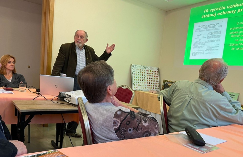 Fotografia zachytáva interiér malej konferenčnej miestnosti, kde prebieha odborná prednáška. V ľavej časti záberu stojí starší muž, Ing. Viliam Stockmann, PhD., ktorý je práve pri svojej prezentácii. Gestom pravou rukou ukazuje na premietnutý obraz na stenu. Má šedivé vlasy, bradu a okuliare, oblečený je v tmavom saku a svetlej košeli so vzorom. Pred ním sa na stole nachádza otvorený laptop a biely projektor, ktorý premieta zelený diapozitív s textom o 70. výročiu uzákonenia štátnej ochrany prírody. Vedľa neho sedí žena s blond vlasmi v čiernom svetri, ktorá pozorne sleduje prezentáciu. Vpravo v popredí sedia starší muž a žena, obaja chrbtom k fotoaparátu. Žena má krátke sivé vlasy a mikinu s výrazným vzorom z kruhov na rukáve. Muž vedľa nej má svetlo zelenú košeľu. Na stole pred nimi leží diaľkový ovládač projektora a papierové materiály. Vedľa premietanej plochy sú na stole vystavené farebné informačné tabule s obrázkami prírody. Celková atmosféra fotografie je profesionálna a sústredená, zachytávajúca moment počas formálnej prednášky venovanej ochrane prírody.