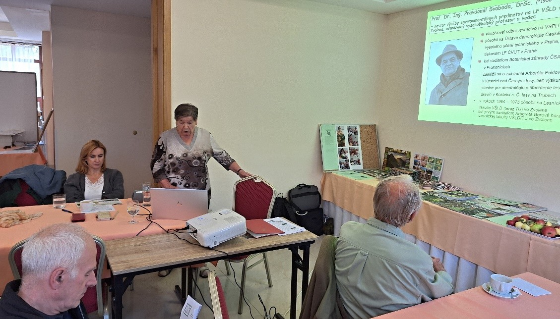 Fotografia zachytáva scénu z malej konferenčnej miestnosti počas odborného stretnutia. V strede záberu stojí staršia žena, Doc. Ing. Margita Jančová, PhD., ktorá práve prednáša. Má krátke sivé vlasy a oblečenú svetlú blúzku s hnedo-sivým vzorom. Pravou rukou sa opiera o operadlo stoličky, pred ňou je otvorený laptop a biely projektor položený na stole. Na ľavej strane sedí mladšia žena s blond vlasmi v tmavom svetri a bielej blúzke, ktorá ju pozorne počúva. V popredí sú dvaja starší muži sediaci chrbtom k fotoaparátu, sledujúci premietanú prezentáciu na stene. Na stene sa premieta diapozitív so zeleným pozadím, textom a fotografiou muža v klobúku. Na stole vpravo sú rozložené rôzne informačné materiály a miska s jablkami. Celková atmosféra miestnosti je pokojná a sústredená, typická pre formálne odborné podujatie.