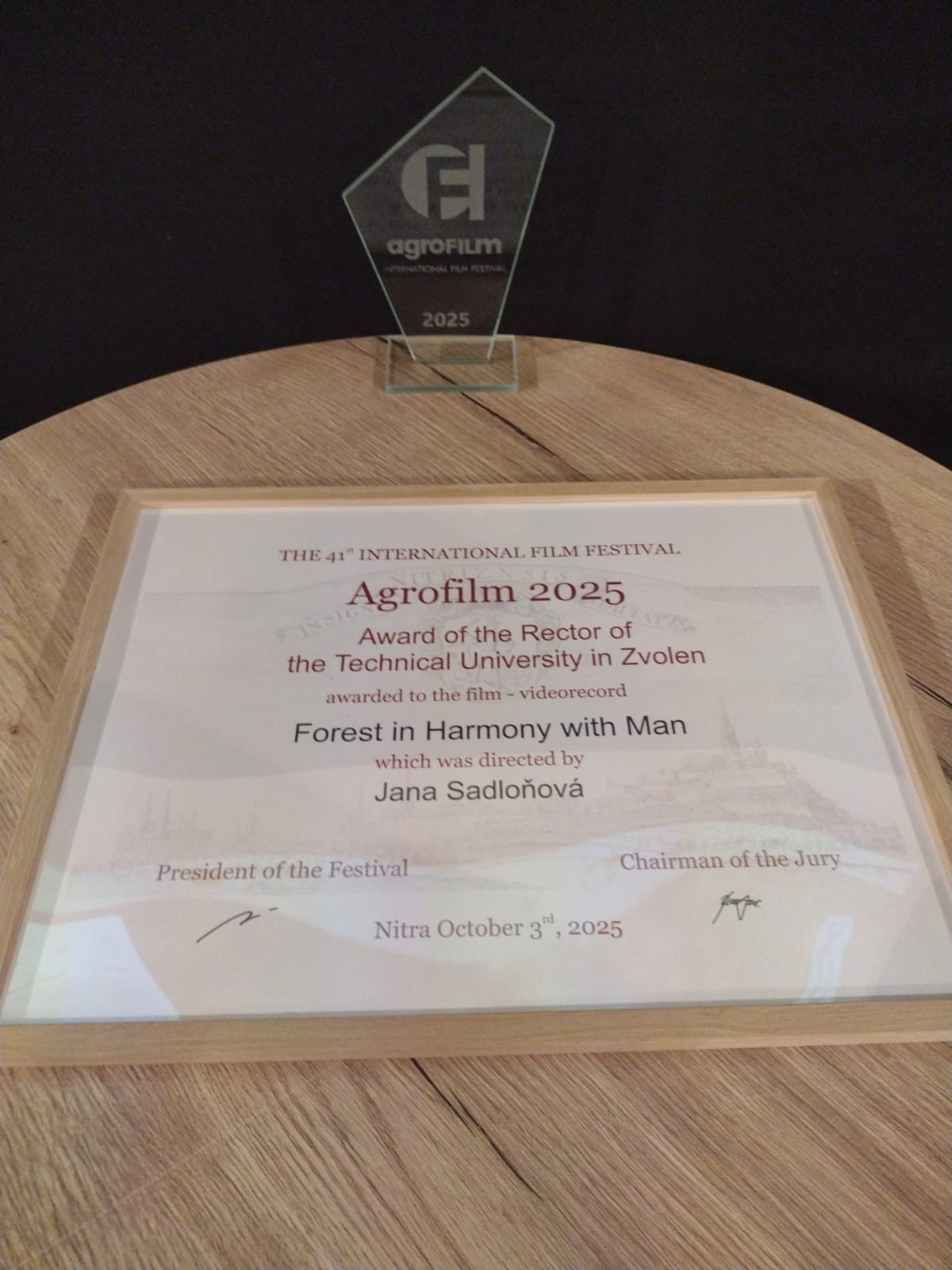 Na fotografii je zobrazené ocenenie udelené filmu Forest in Harmony with Man (slovensky Les v harmónii s človekom) na 41. ročníku medzinárodného filmového festivalu Agrofilm 2025. Ocenenie pozostáva z: Sklenenej trofeje v tvare geometrického päťuholníka s logom Agrofilm a nápisom „Agrofilm International Film Festival 2025“. Diplomu v drevenom ráme, ktorý obsahuje anglický text: Agrofilm 2025 – Award of the Rector of the Technical University in Zvolen, ocenenie bolo udelené filmu Forest in Harmony with Man, réžia: Jana Sadloňová. Dátum: Nitra, 3. októbra 2025. Podpísané prezidentom festivalu a predsedom poroty. Ocenenie je umiestnené na okrúhlom drevenom stole pred čiernym pozadím.