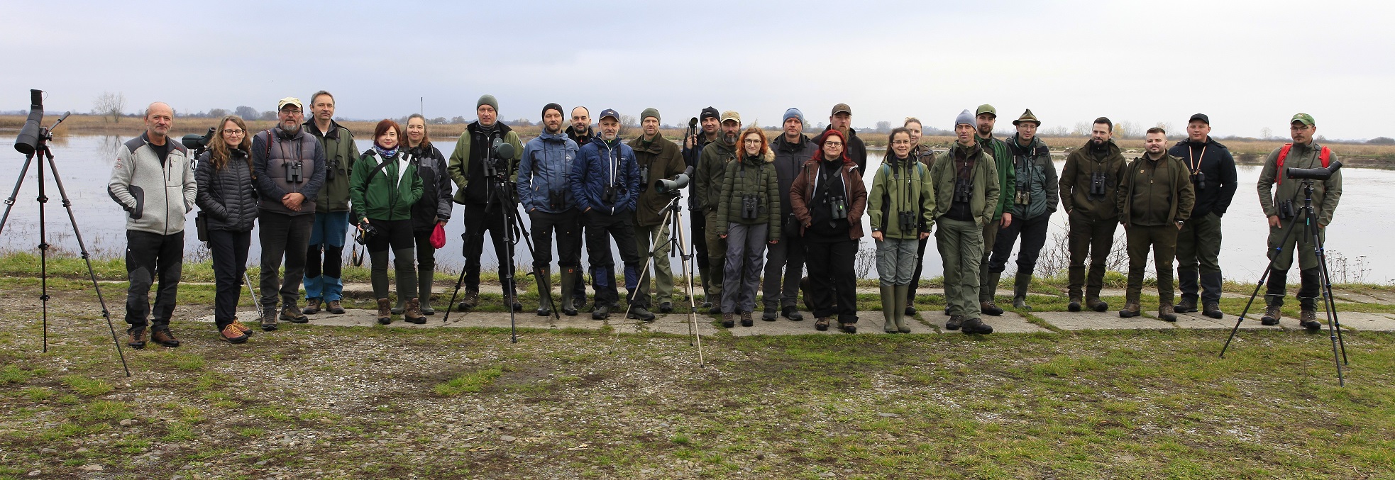 Účastníci školenia zameraného na monitoring zimujúceho vodného vtáctva počas terénnej časti na Senianskych rybníkoch. Veľká skupina ľudí stojí v rade pred vodnou hladinou, viacerí majú ďalekohľady a statívy na pozorovanie vtáctva. Všetci sú oblečení v outdoorovom oblečení do chladného počasia.