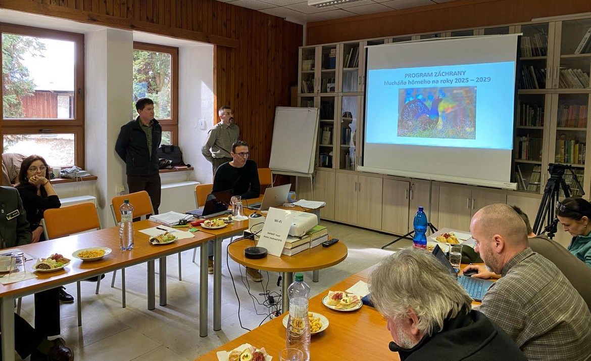 Skupina ľudí sedí okolo konferenčného stola počas pracovného stretnutia v zasadačke Správy Národného parku Nízke Tatry. Na stole sú dokumenty, občerstvenie a fľaše s vodou. V pozadí je baner s logom parku a fotografiou horskej krajiny.