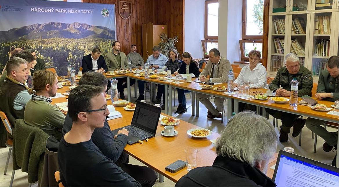 Skupina ľudí sedí okolo konferenčného stola počas pracovného stretnutia v zasadačke Správy Národného parku Nízke Tatry. Na stole sú dokumenty, občerstvenie a fľaše s vodou. V pozadí je baner s logom parku a fotografiou horskej krajiny.