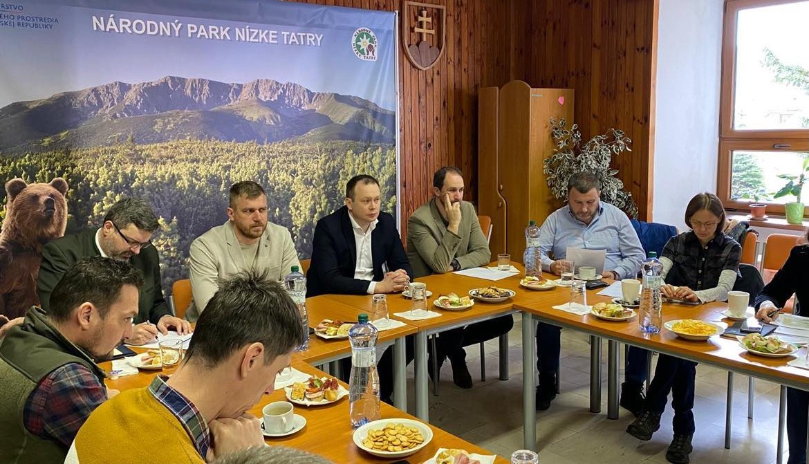 Skupina ľudí sedí okolo konferenčného stola počas pracovného stretnutia v zasadačke Správy Národného parku Nízke Tatry. Na stole sú dokumenty, občerstvenie a fľaše s vodou. V pozadí je baner s logom parku a fotografiou horskej krajiny.