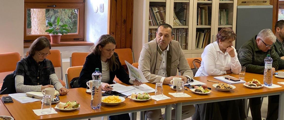 Skupina ľudí sedí okolo konferenčného stola počas pracovného stretnutia v zasadačke Správy Národného parku Nízke Tatry. Na stole sú dokumenty, občerstvenie a fľaše s vodou. V pozadí je baner s logom parku a fotografiou horskej krajiny.