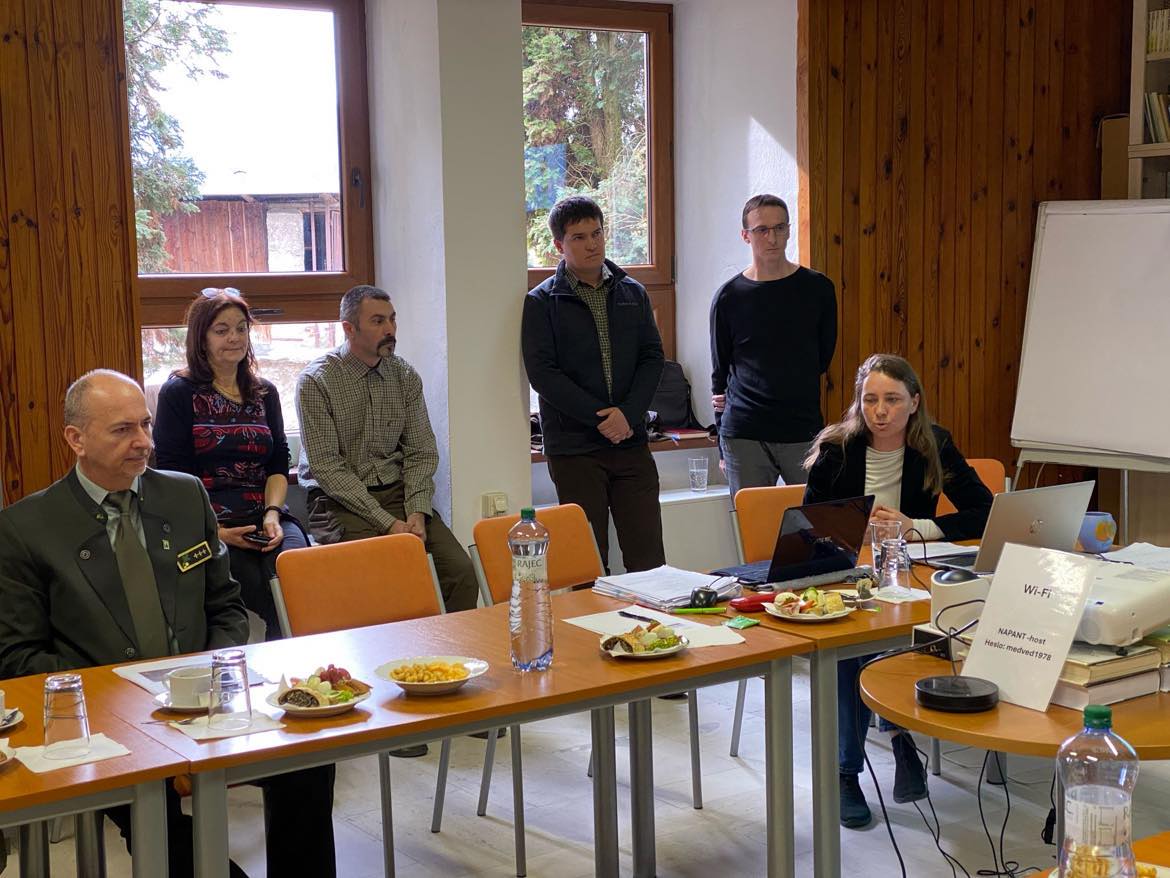 Skupina ľudí sedí okolo konferenčného stola počas pracovného stretnutia v zasadačke Správy Národného parku Nízke Tatry. Na stole sú dokumenty, občerstvenie a fľaše s vodou. V pozadí je baner s logom parku a fotografiou horskej krajiny.