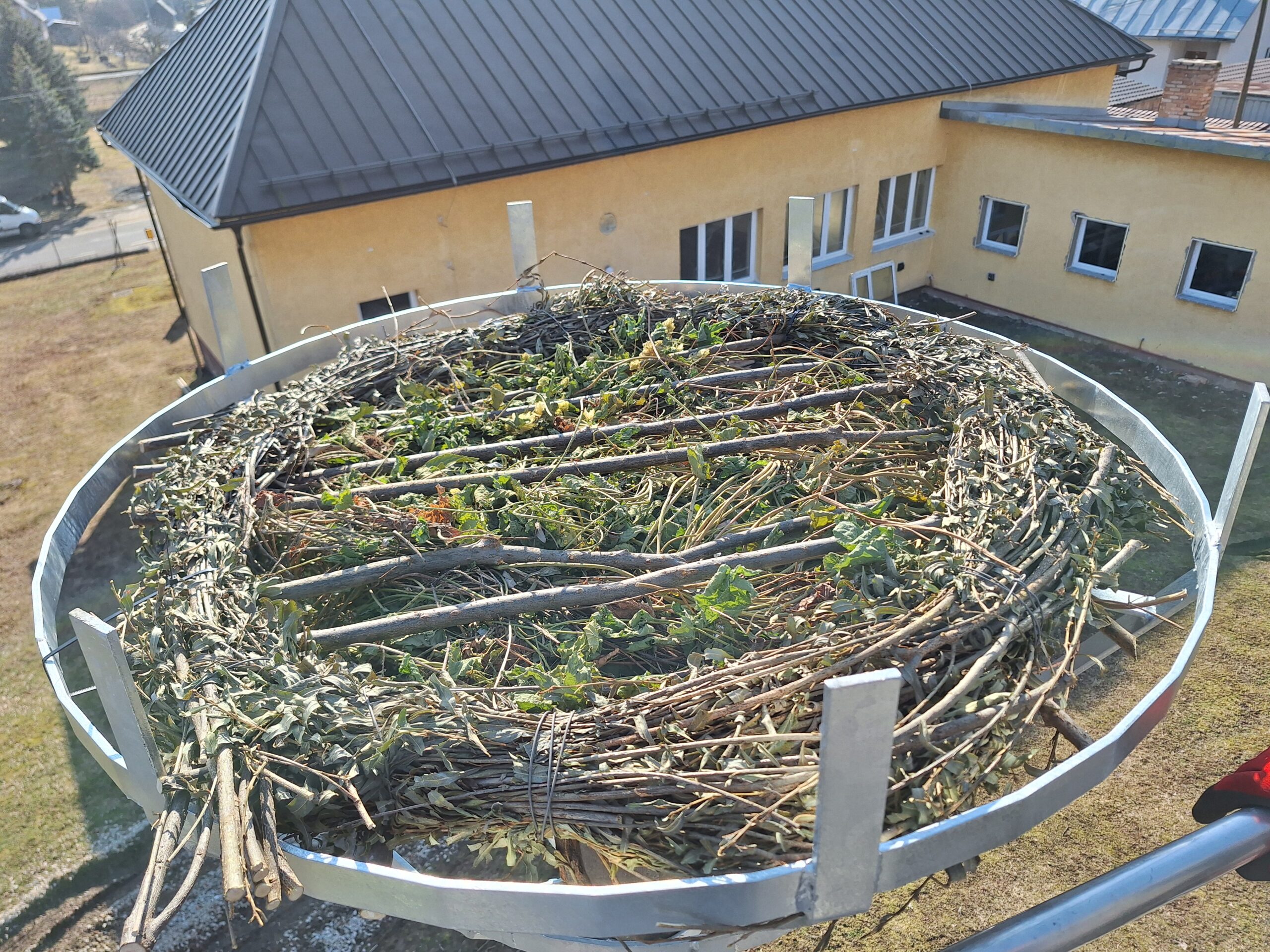 Detail novovybudovanej kovovej podložky pre bocianie hniezdo, naplnenej vetvami a zeleňou. Podložka je umiestnená na stĺpe vo výške, v pozadí je budova s plechovou strechou a niekoľko okien.