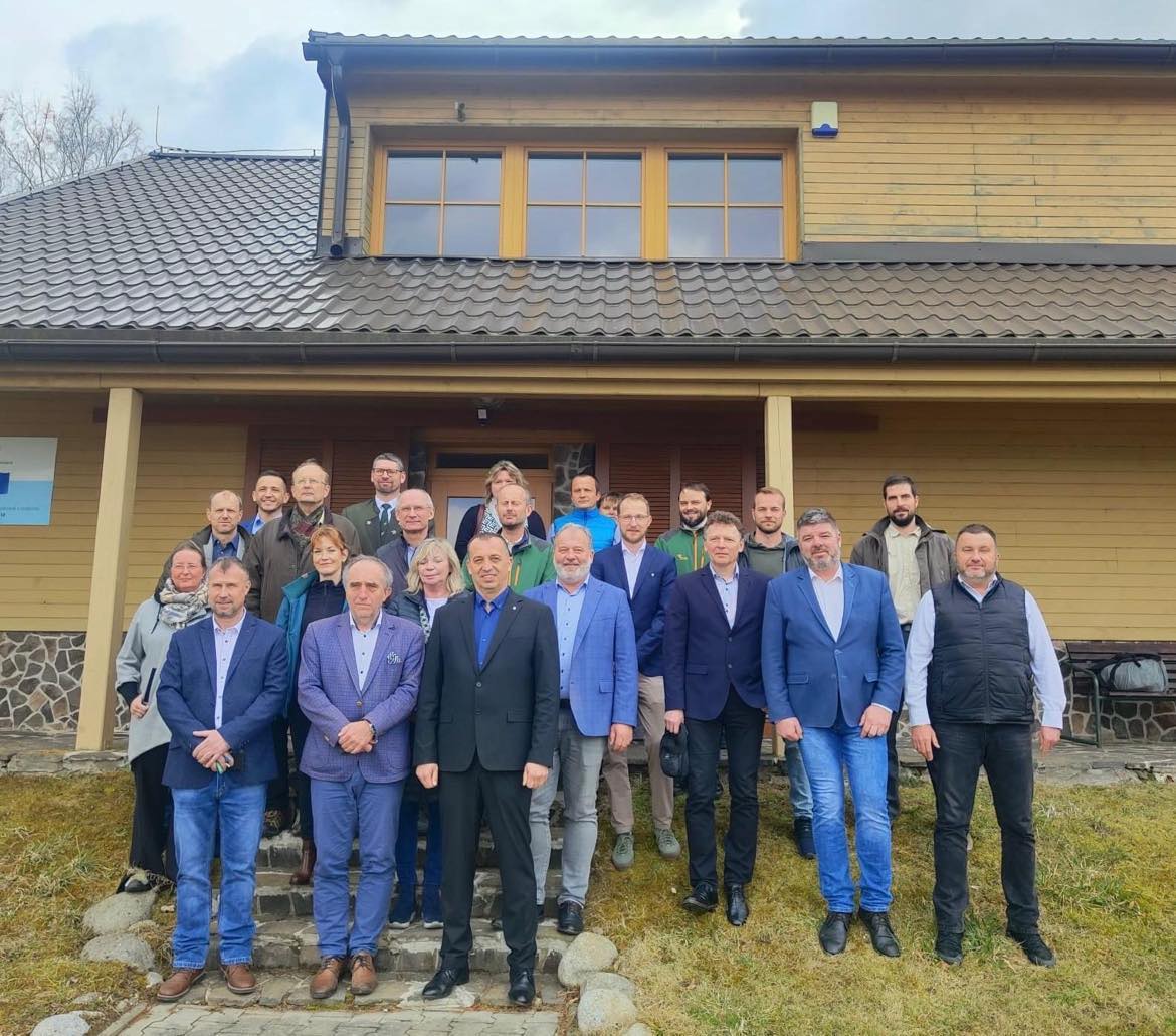 Skupinová fotografia účastníkov stretnutia stojacich pred drevenou budovou s kamenným základom. Ide o spoločnú snímku viacerých zástupcov organizácií a odborníkov z oblasti ochrany prírody.