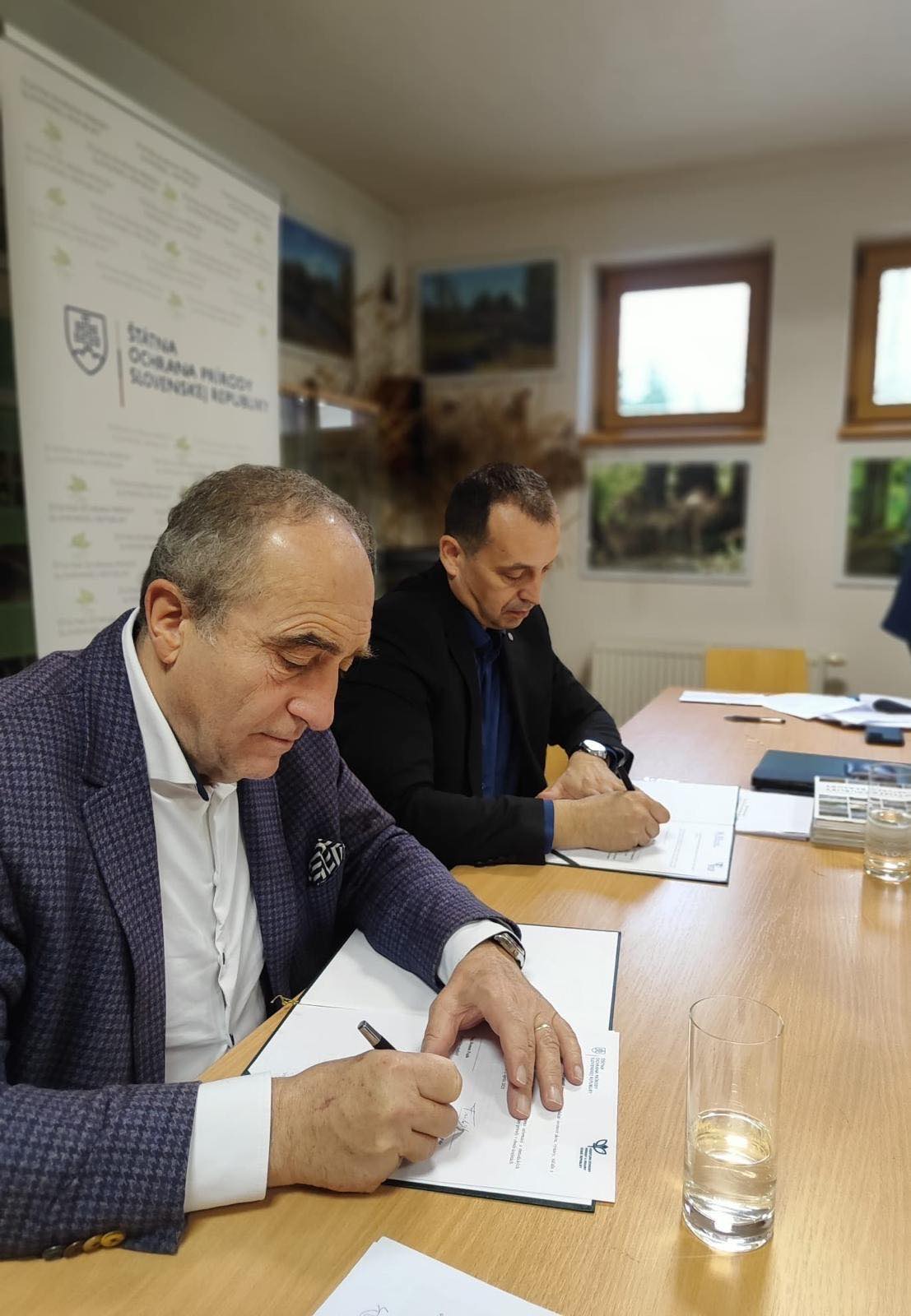 Dvaja muži sedia za stolom a podpisujú dokumenty počas oficiálneho rokovania v zasadačke Štátnej ochrany prírody Slovenskej republiky. Na stole sú pripravené zmluvy, perá a poháre s vodou, v pozadí vidno informačný banner ŠOP SR.