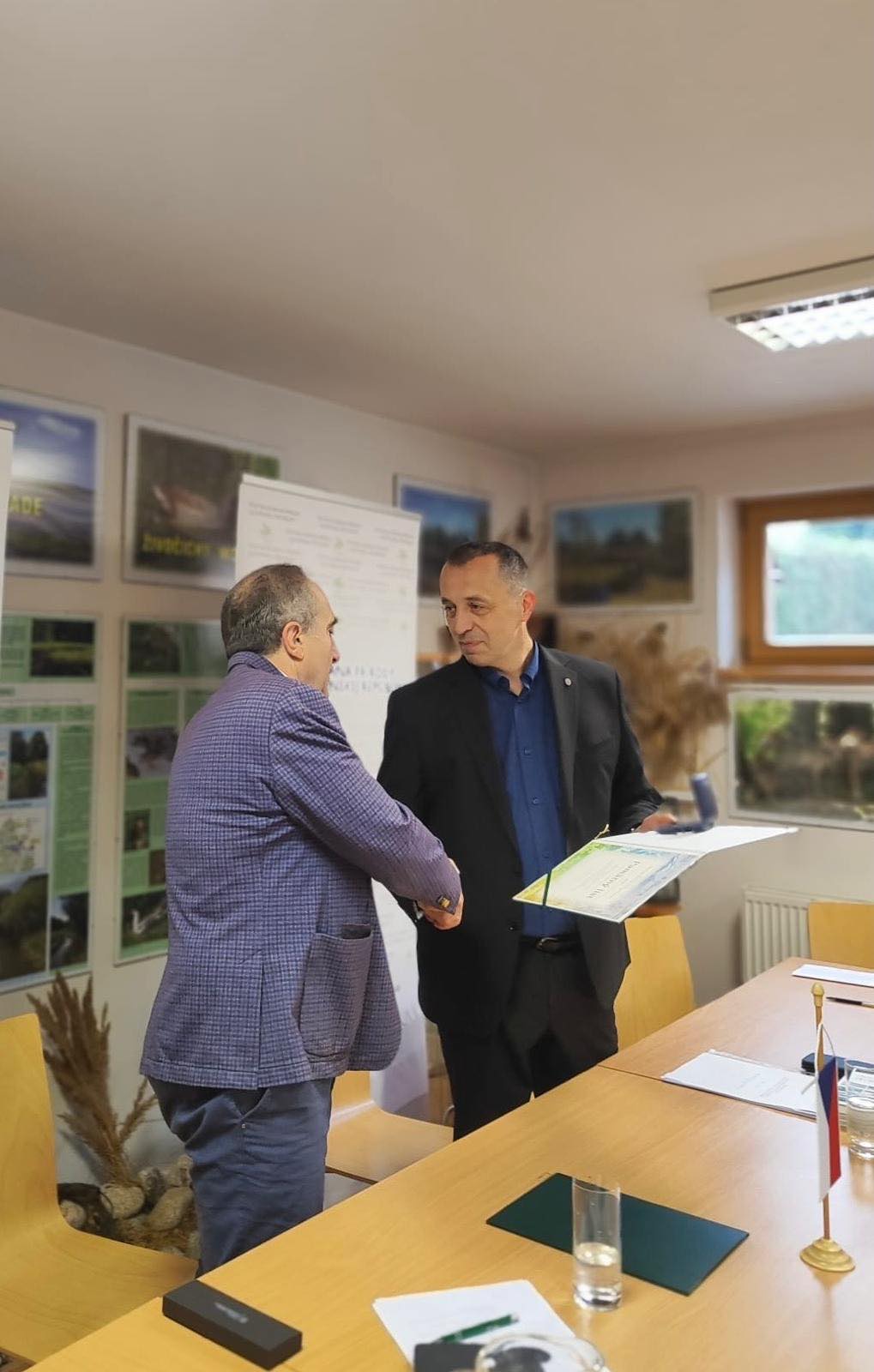 Dvaja muži si podávajú ruky pri oficiálnom akte podpisu dokumentu v zasadačke Štátnej ochrany prírody Slovenskej republiky. Jeden z nich drží v ruke zložku s podpísanou dohodou.