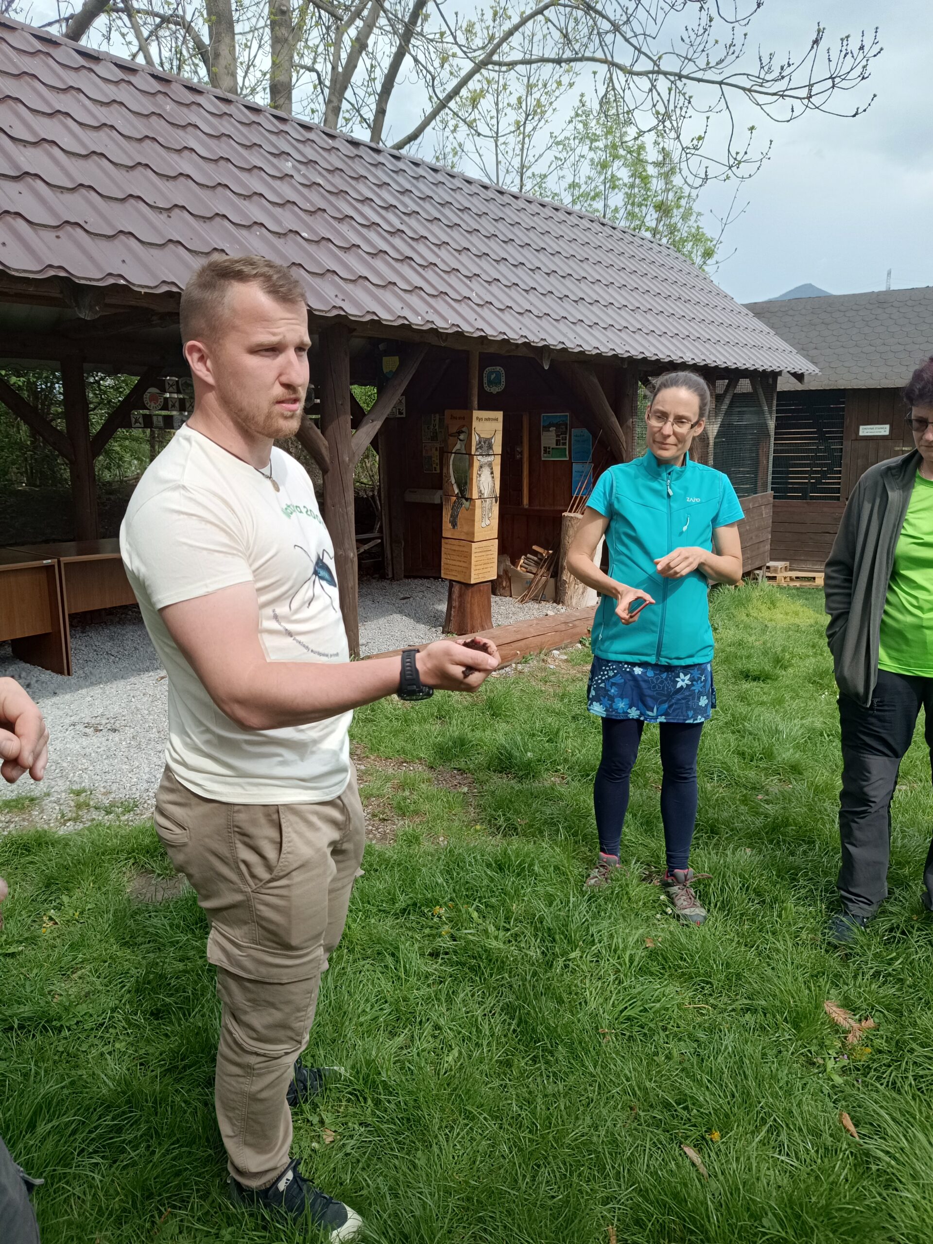 Muž v béžovom tričku stojí na tráve a ukazuje skupine ľudí drobný predmet, pravdepodobne prírodný materiál. Ostatní účastníci ho pozorne sledujú. V pozadí je drevený altánok s informačnými panelmi o vtákoch.