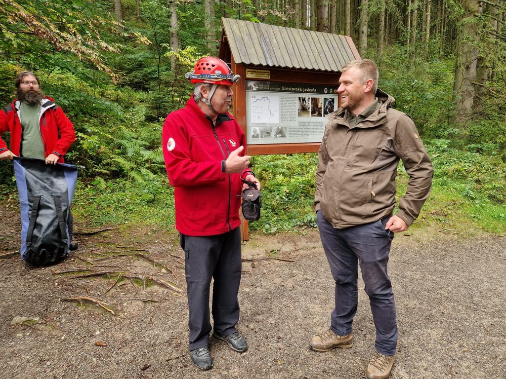 Pracovník Správy slovenských jaskýň v červenej prilbe diskutuje s účastníkom podujatia pri informačnom paneli Brestovskej jaskyne v lesnom prostredí.