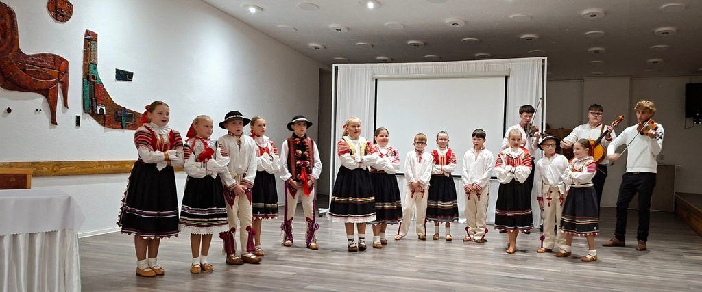 Detský folklórny súbor v tradičných krojoch vystupuje v interiéri, v sprievode hudobníkov s husľami a violončelom.